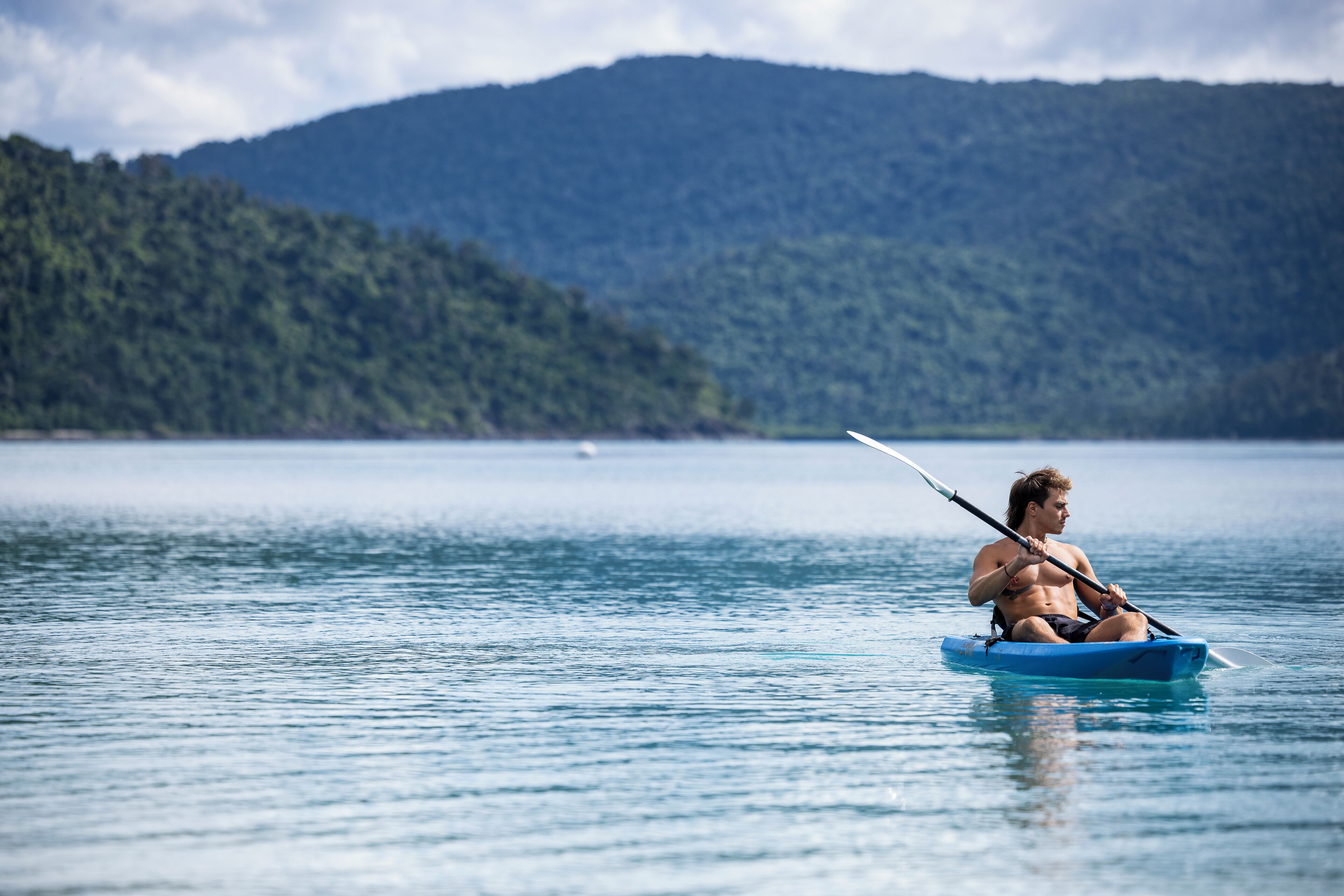 Une personne dans un kayak bleu sur un lac calme, avec une montagne boisée en arrière-plan.