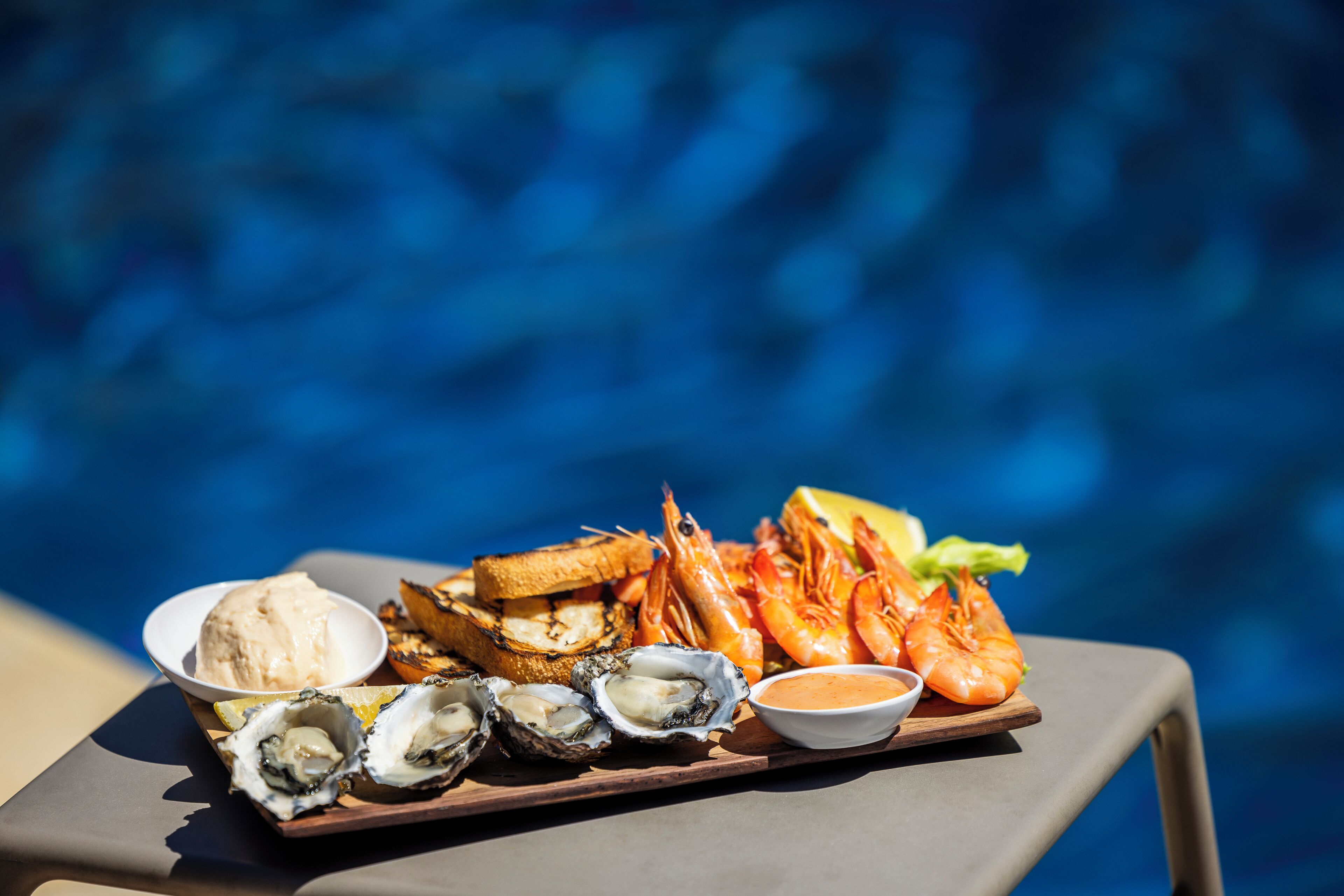 A tray of seafood including shrimp, oysters, and grilled bread by a pool.