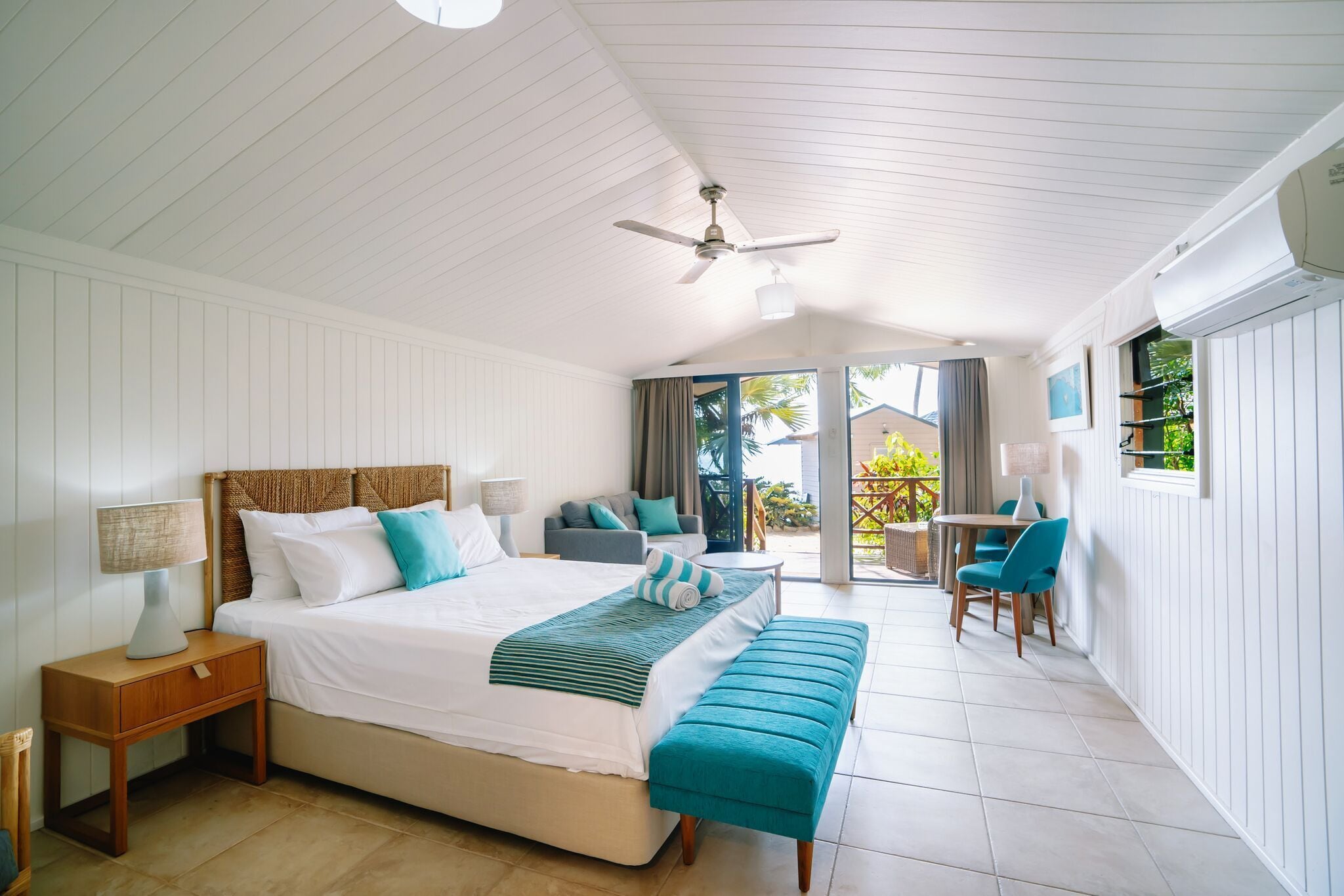 A bedroom with a bed, bedside tables, a bench, a ceiling fan, and a view of the outside.
