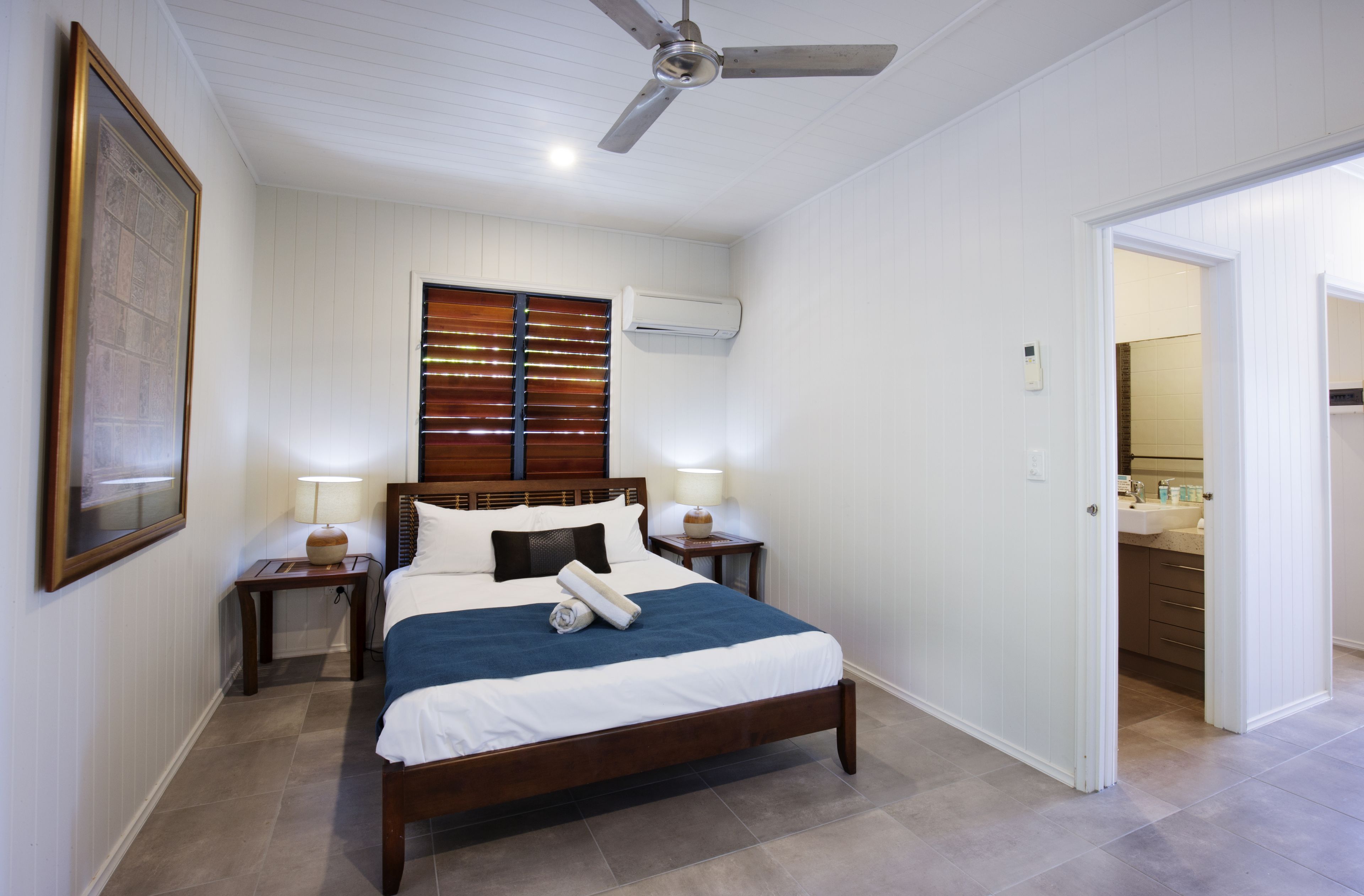 A bedroom with a bed, two bedside tables, a ceiling fan, a window with blinds, and an air conditioning unit.