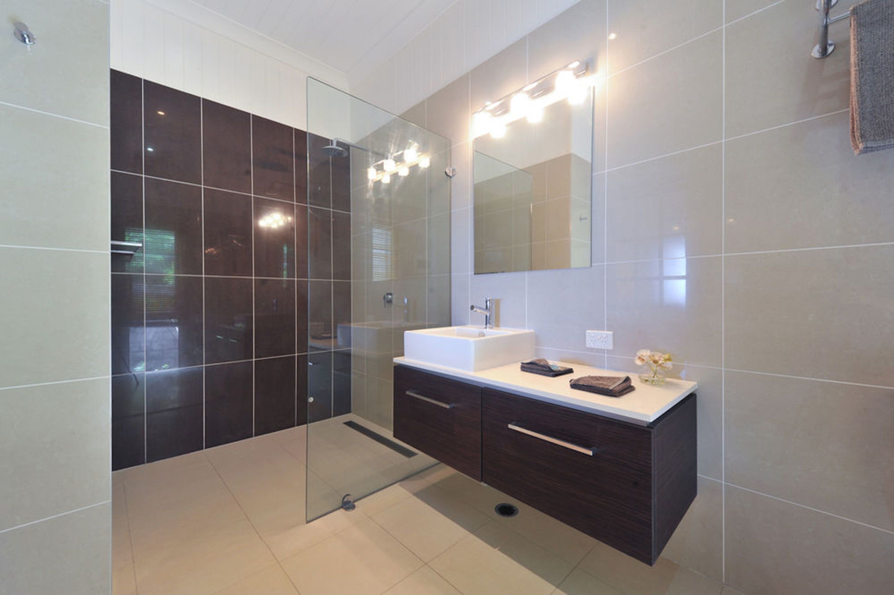 A modern bathroom with a glass-enclosed shower, a white sink, dark cabinetry, and a large mirror with lights above it.