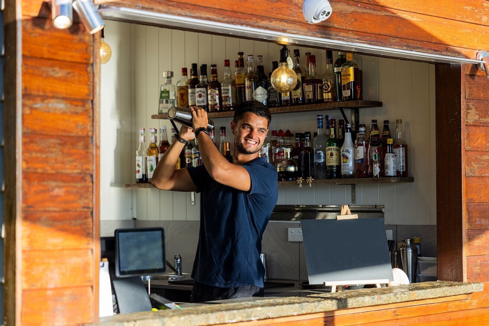 En bartender laver en drink i en hyggelig bar med hylder fyldt med forskellige flasker spiritus.