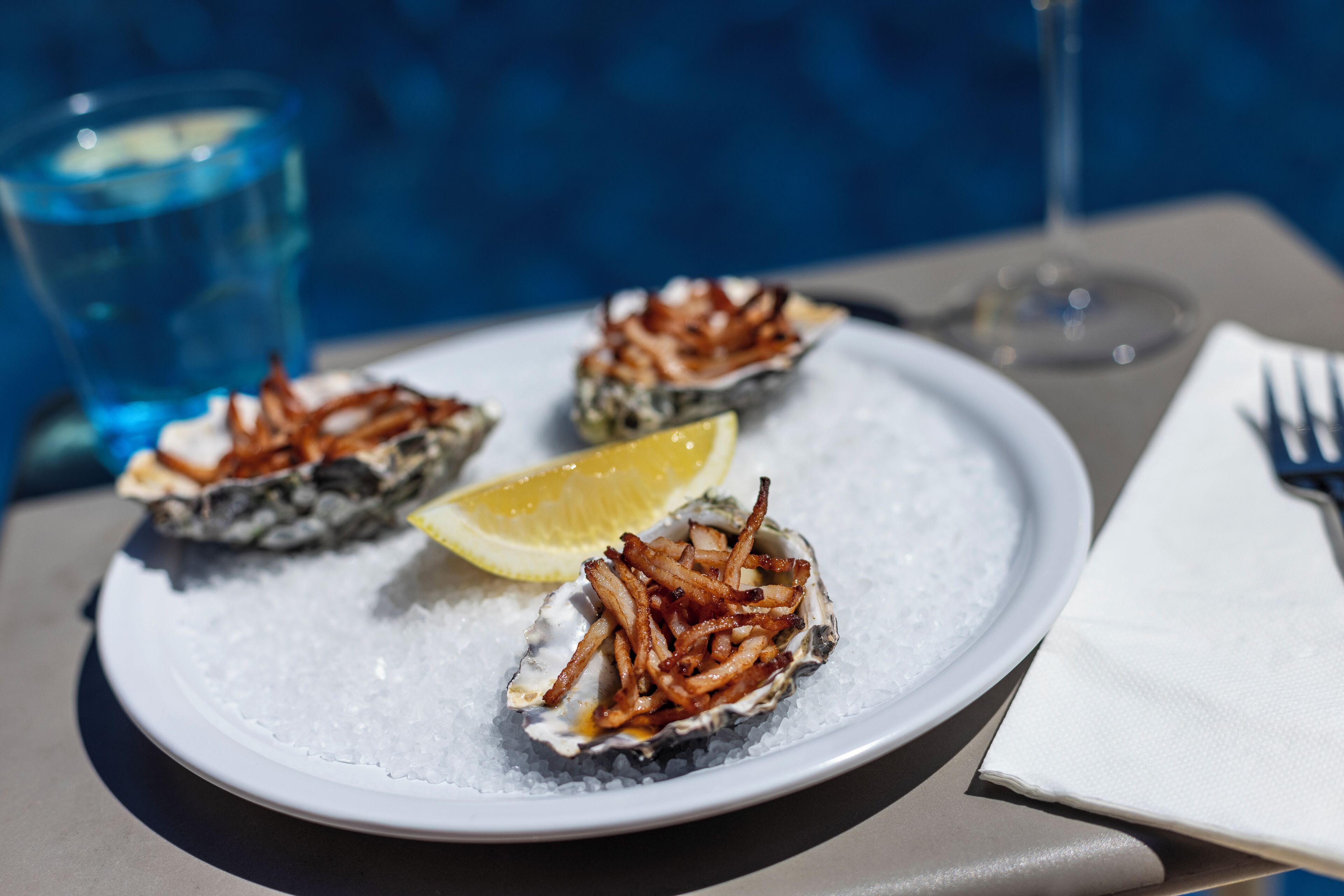 A plate of oysters with lemon wedges and fried onions on a bed of ice, accompanied by a glass of water and a wine glass.