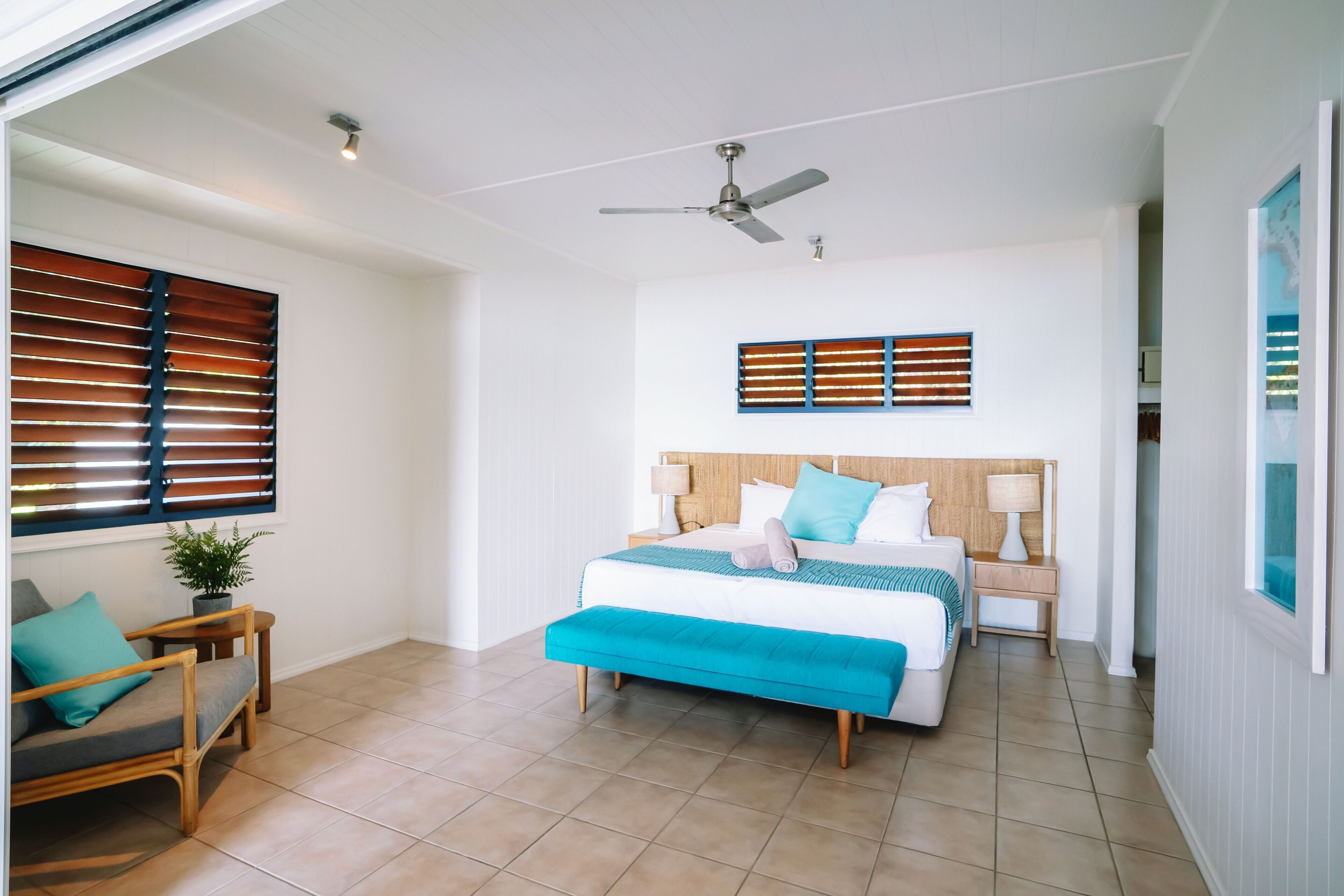 A bedroom with a bed, a ceiling fan, a window with blinds, a bedside table with a lamp, and a small plant.