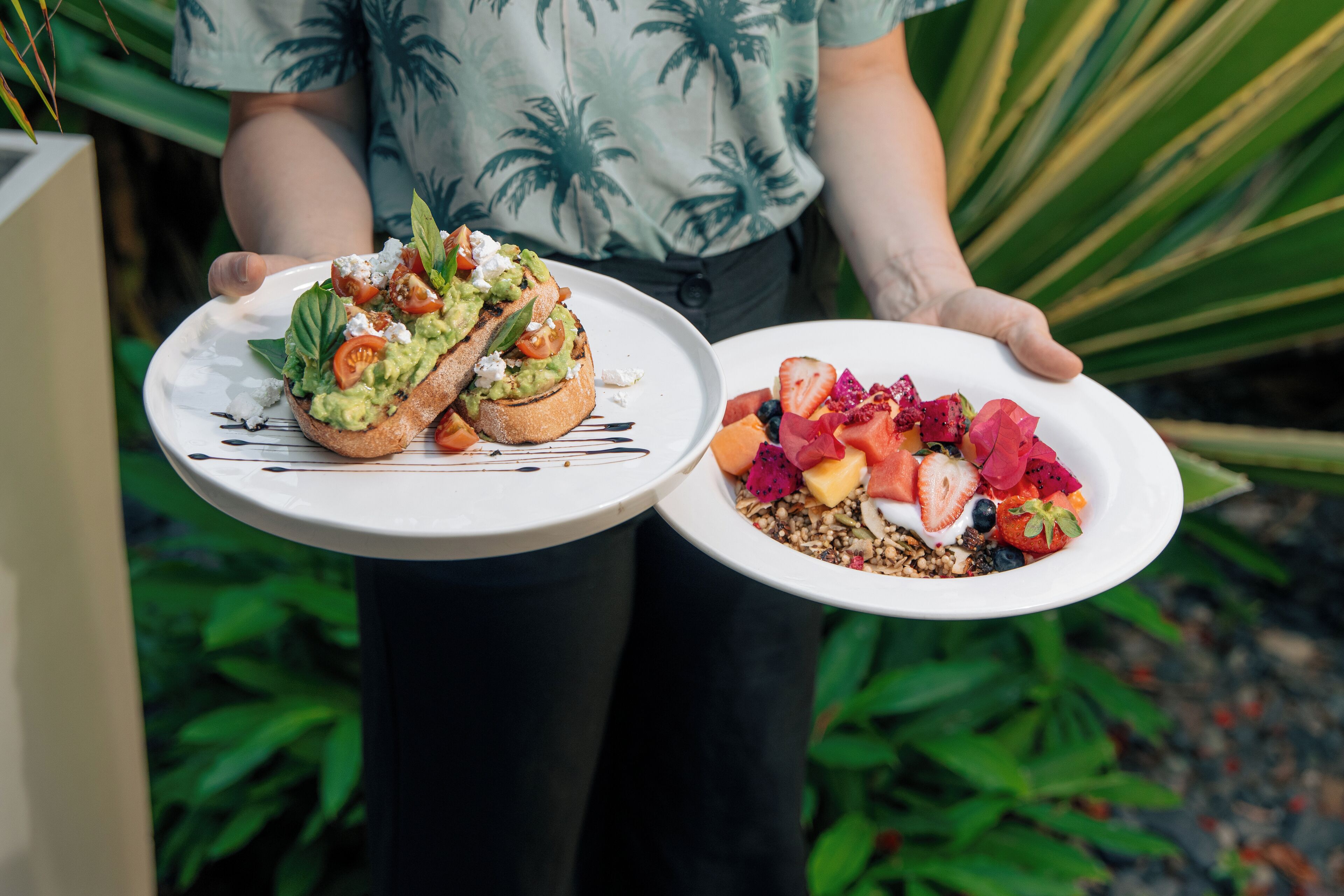 A person holding two plates of food, one with a sandwich and the other with a fruit salad.
