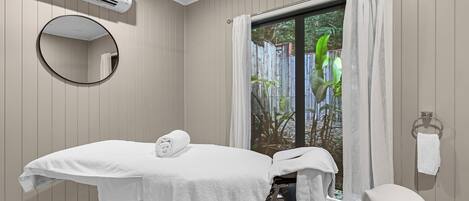 A spa room with a massage table, a large mirror, a window with curtains, and an air conditioning unit.