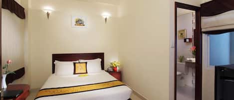 A hotel room with a bed, bedside table, and a small desk with a chair. There is a bathroom visible through an open door.