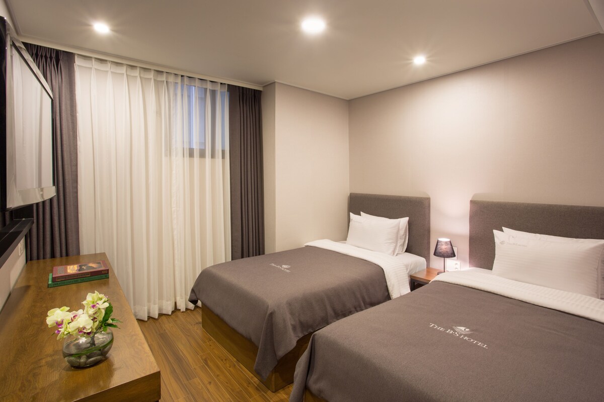 Une chambre d’hôtel avec deux lits, un bureau en bois, un vase avec des fleurs et un livre.