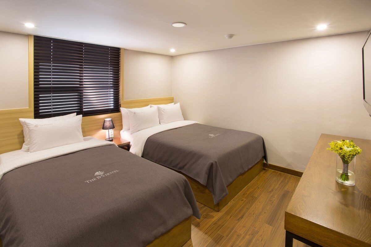 Une chambre d’hôtel avec deux lits, une tête de lit en bois, une table de chevet avec une lampe et une table en bois ornée d’un vase de fleurs.