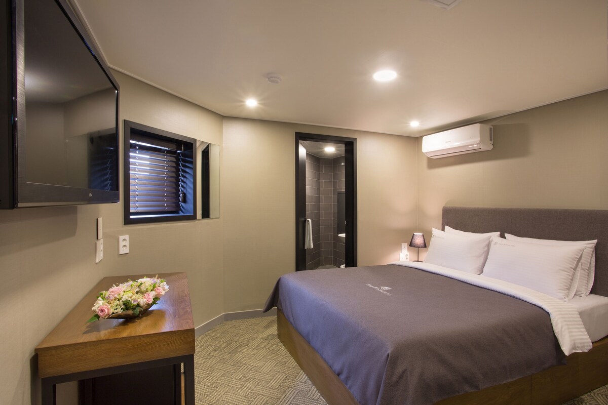 Une chambre d’hôtel avec un lit, un téléviseur fixé au mur, une salle de bain avec douche et une petite table sur laquelle est posé un vase de fleurs.