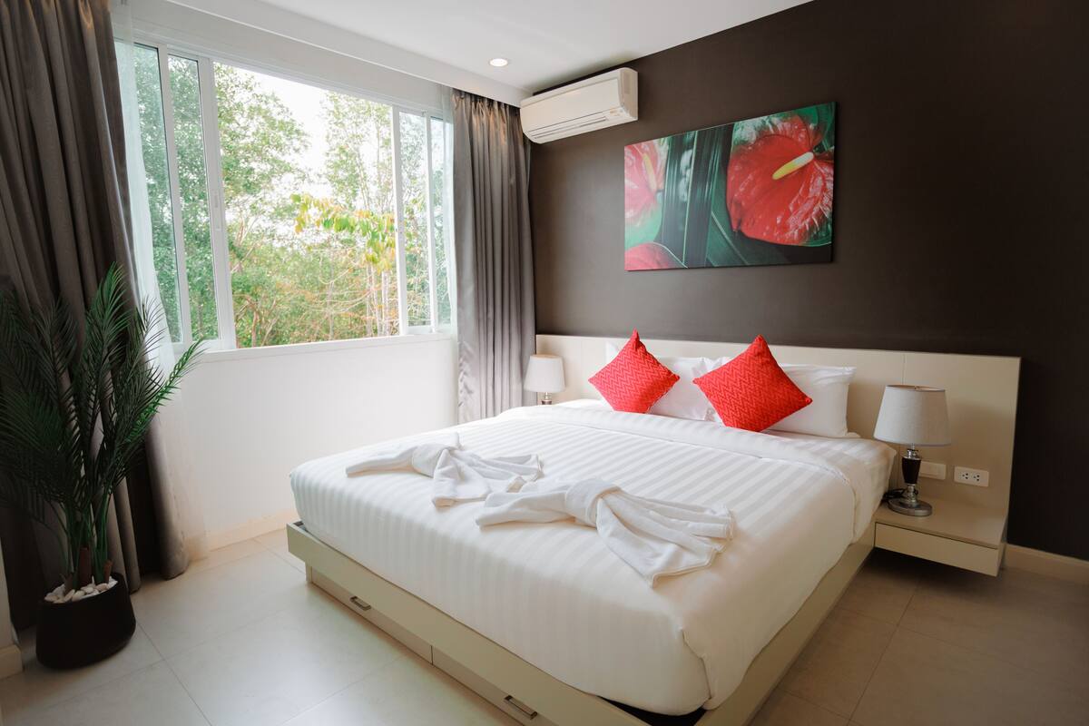 A hotel room with a large bed, a potted plant, a window with curtains, a wall-mounted air conditioner, and a painting.