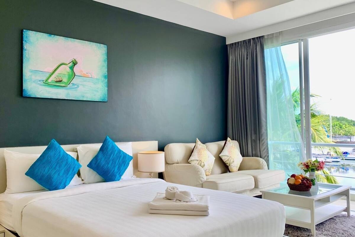 A hotel room with a bed, two side tables, a sofa, and a large window with a view of boats.