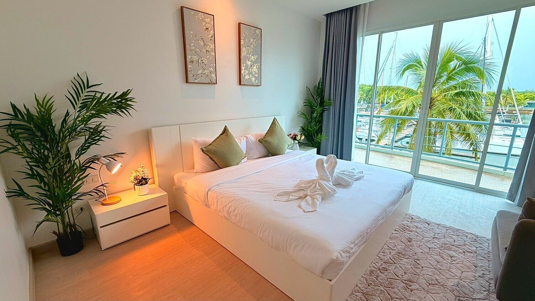 A hotel room with a large bed, a nightstand with a lamp, a potted plant, and a view of palm trees through the window.