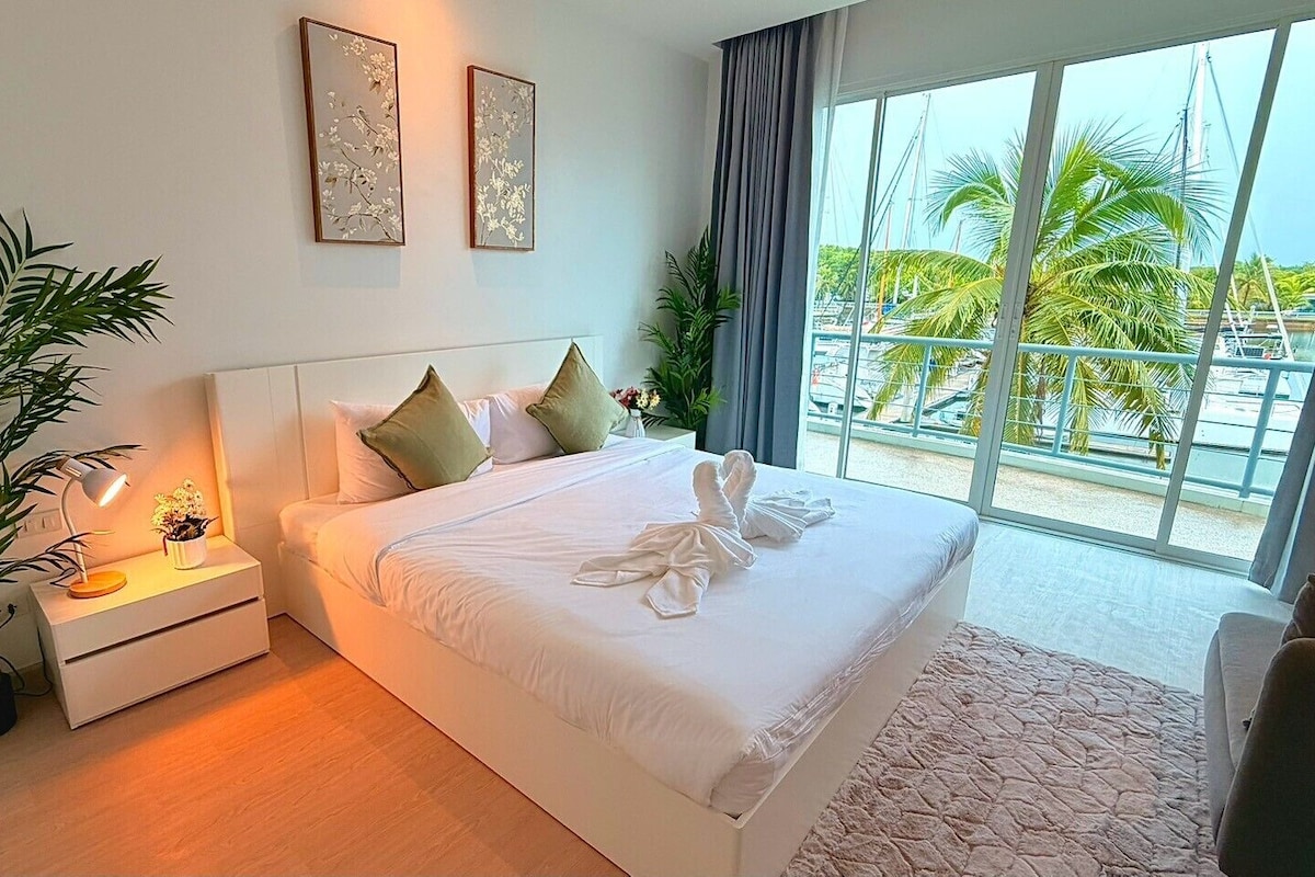 Une chambre d’hôtel avec un grand lit, une table de chevet avec une lampe, une plante en pot et une vue sur les palmiers par la fenêtre.