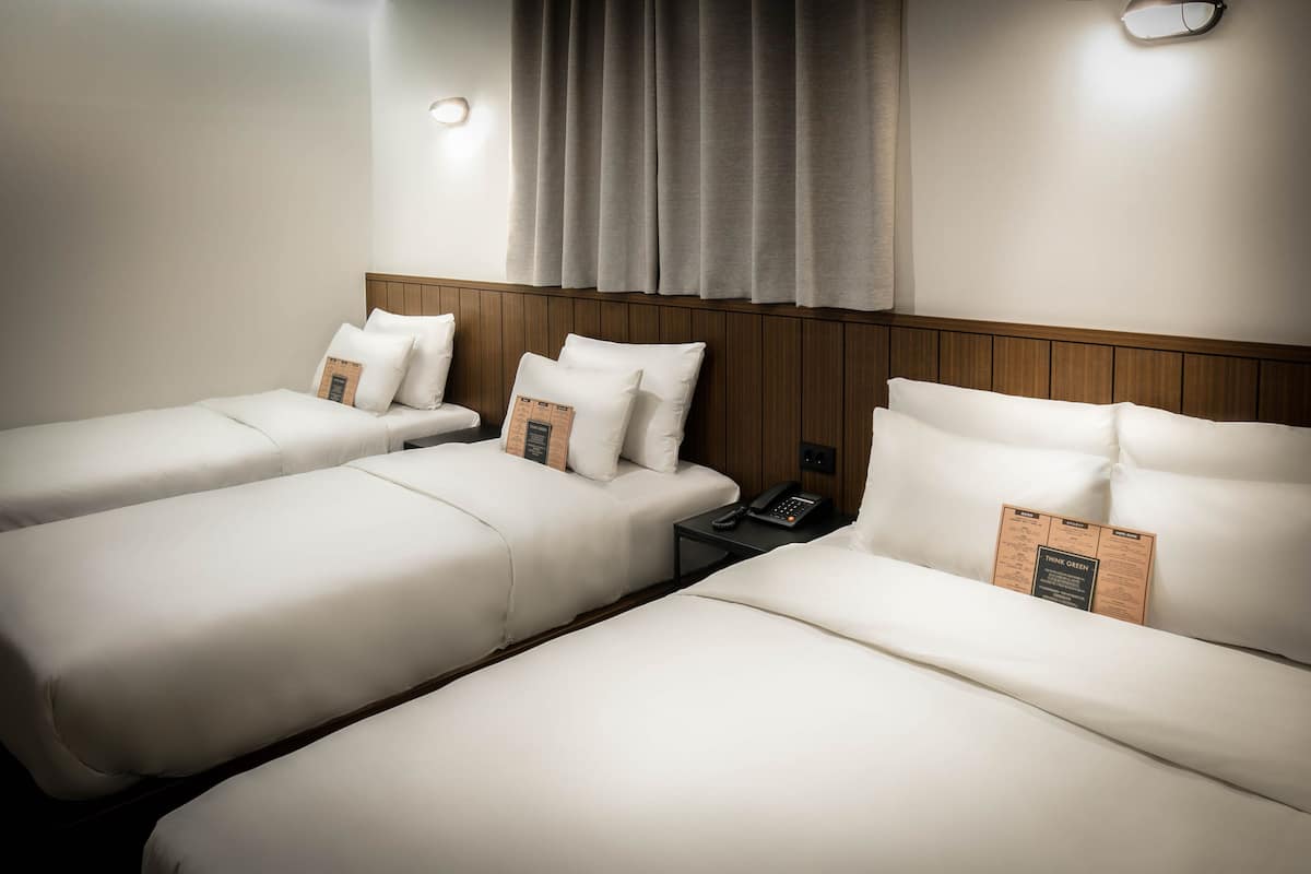 Een hotelkamer met drie enkele bedden, een houten hoofdbord, een nachtkastje met telefoon en een wandlamp.