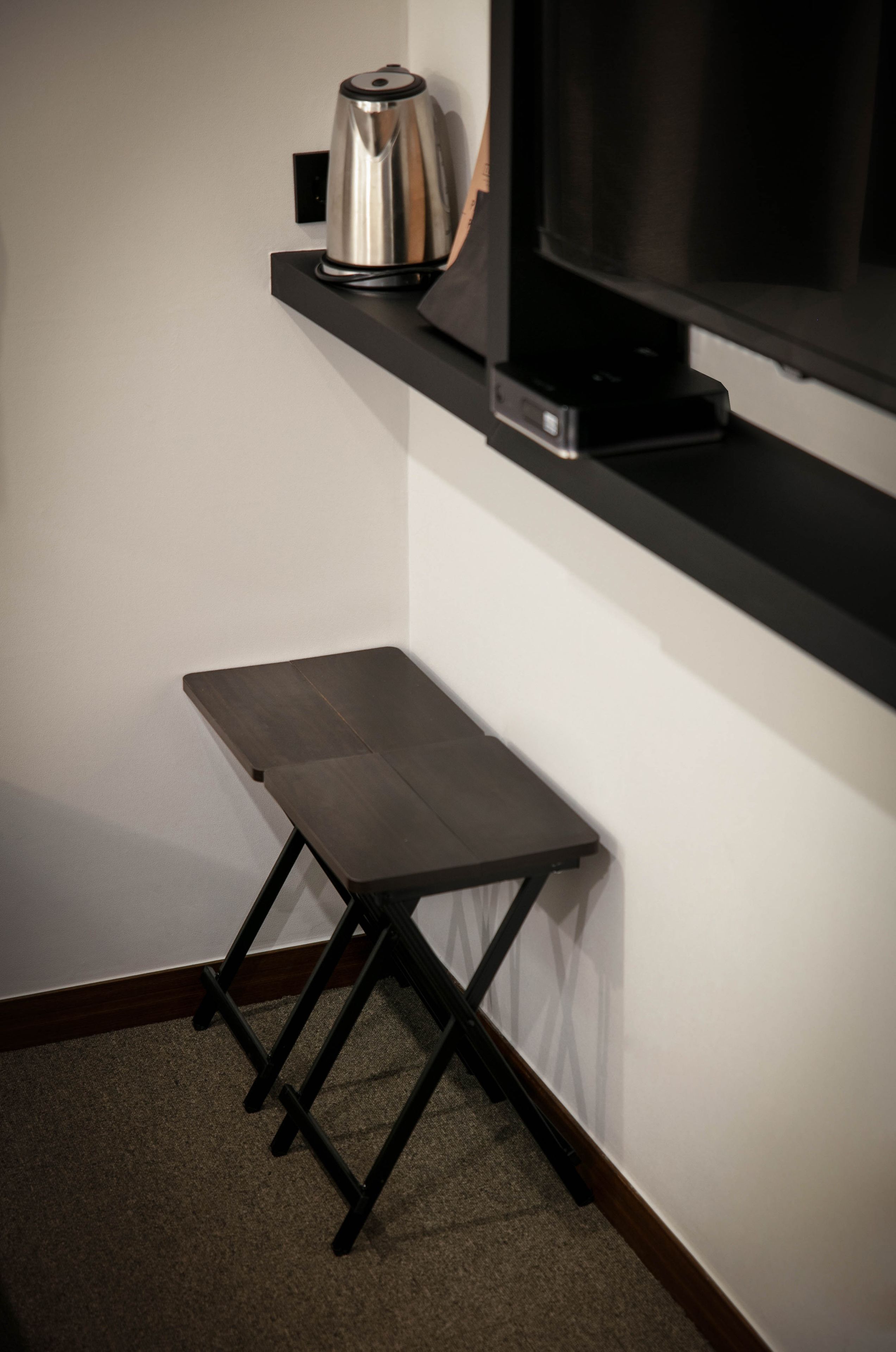 Un taburete pequeño y de madera oscura con una tetera metálica sobre un estante negro, y un televisor montado en la pared.