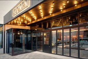 Une entrée d’hôtel moderne avec des portes en verre, une enseigne au-dessus de l’entrée et un intérieur bien éclairé, visible à travers le verre.