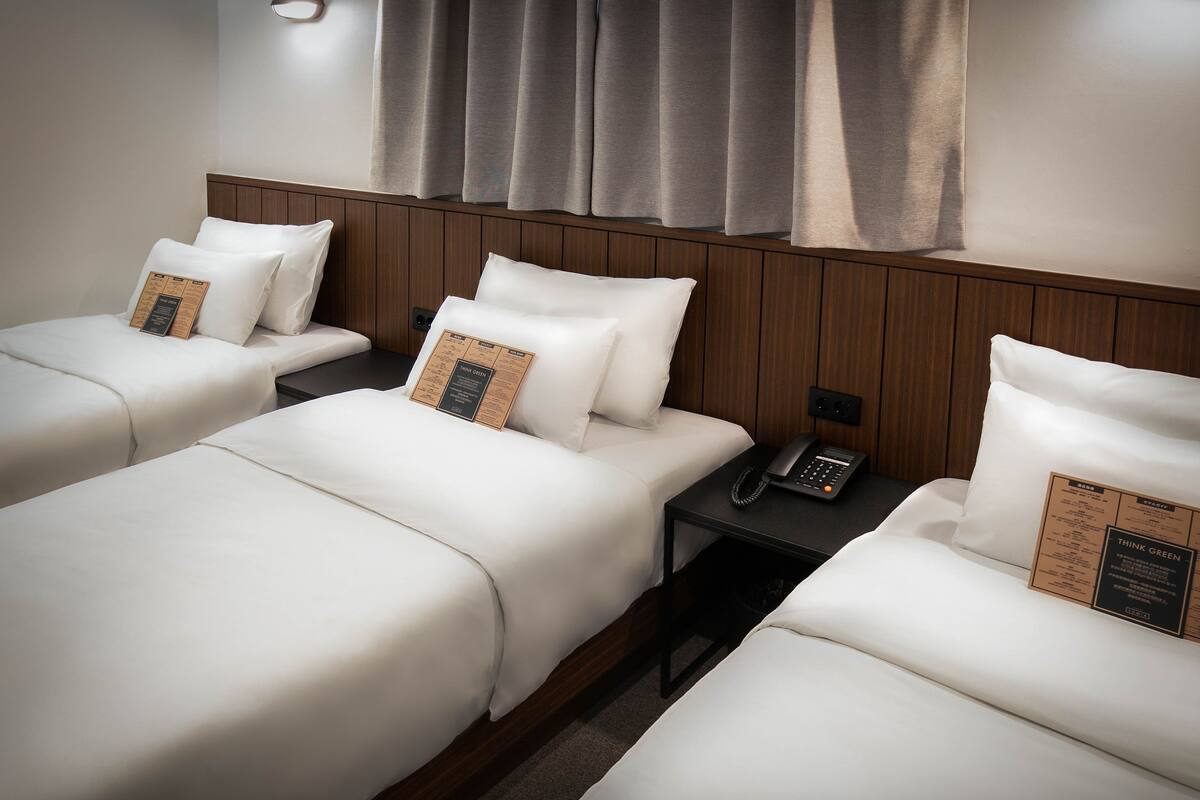 Hotelkamer met drie eenpersoonsbedden, elk voorzien van witte beddengoed en een kussen met een 'Think Green' placemat. Er is een nachtkastje met een zwarte telefoon en een stopcontact.