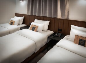 Une chambre d’hôtel avec trois lits simples, chacun doté de draps blancs et d’un oreiller surmonté d’un sous-verre à l’inscription « Pensez vert ». On y trouve une table de chevet avec un téléphone noir et une prise électrique.