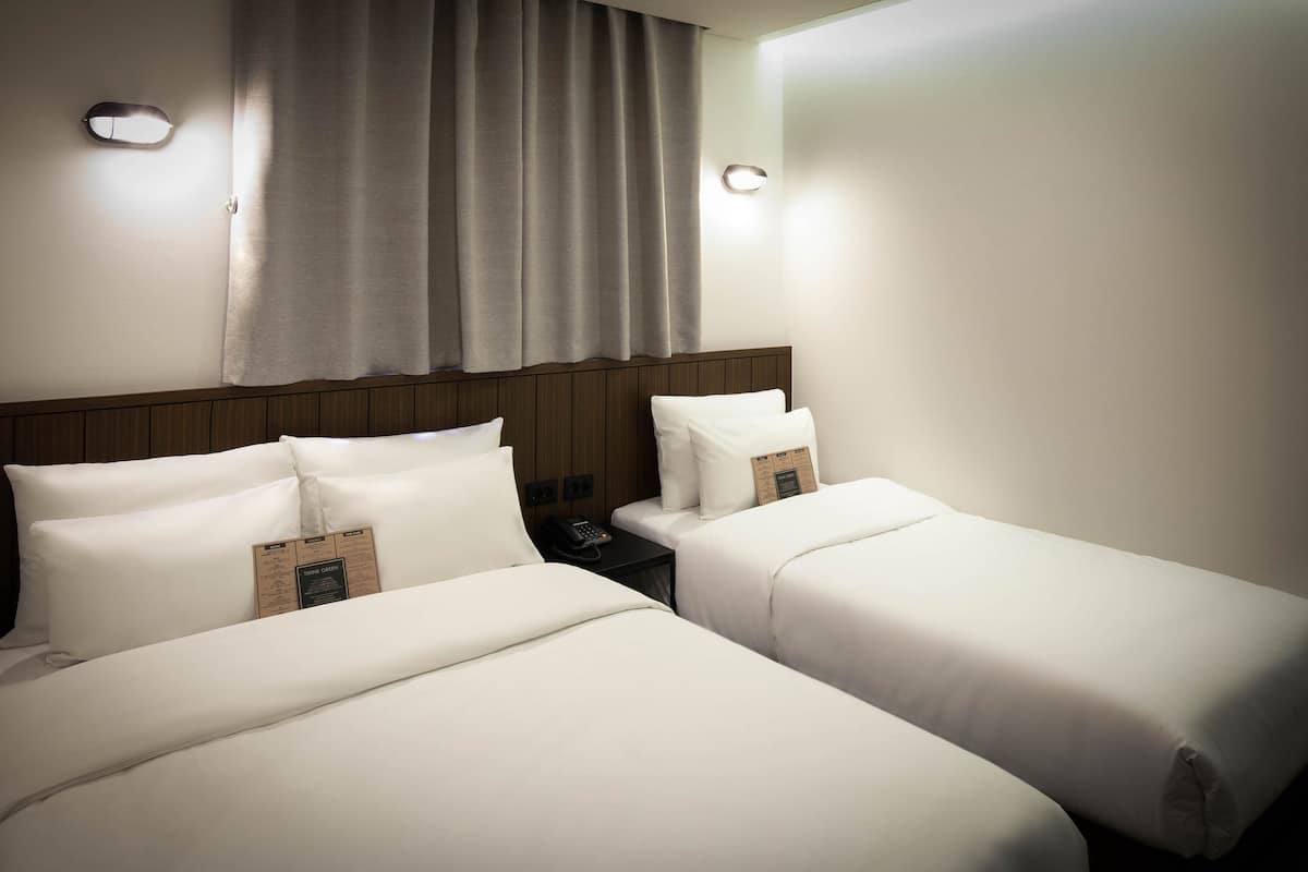 Een hotelkamer met twee bedden, een houten hoofdbord, een nachtkastje met telefoon en een raam met gordijnen.