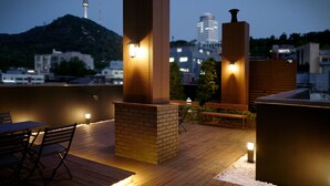 Eine Holzterrasse mit Außenmöbeln und Beleuchtung, mit Blick auf eine Stadtlandschaft mit einem Berg und Gebäuden.