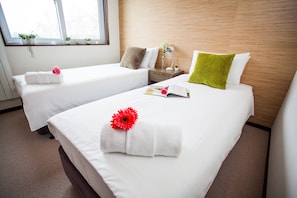 Two neatly made beds with white linens, a small bedside table with a lamp, and a window with plants. - Tamo (Kutchan)