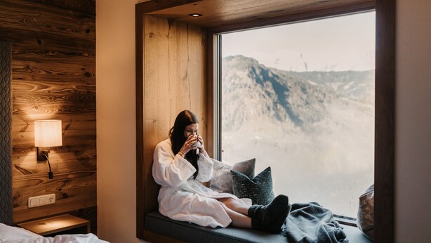 Eine Person sitzt an einem Fenster mit Blick auf eine gebirgige Landschaft.