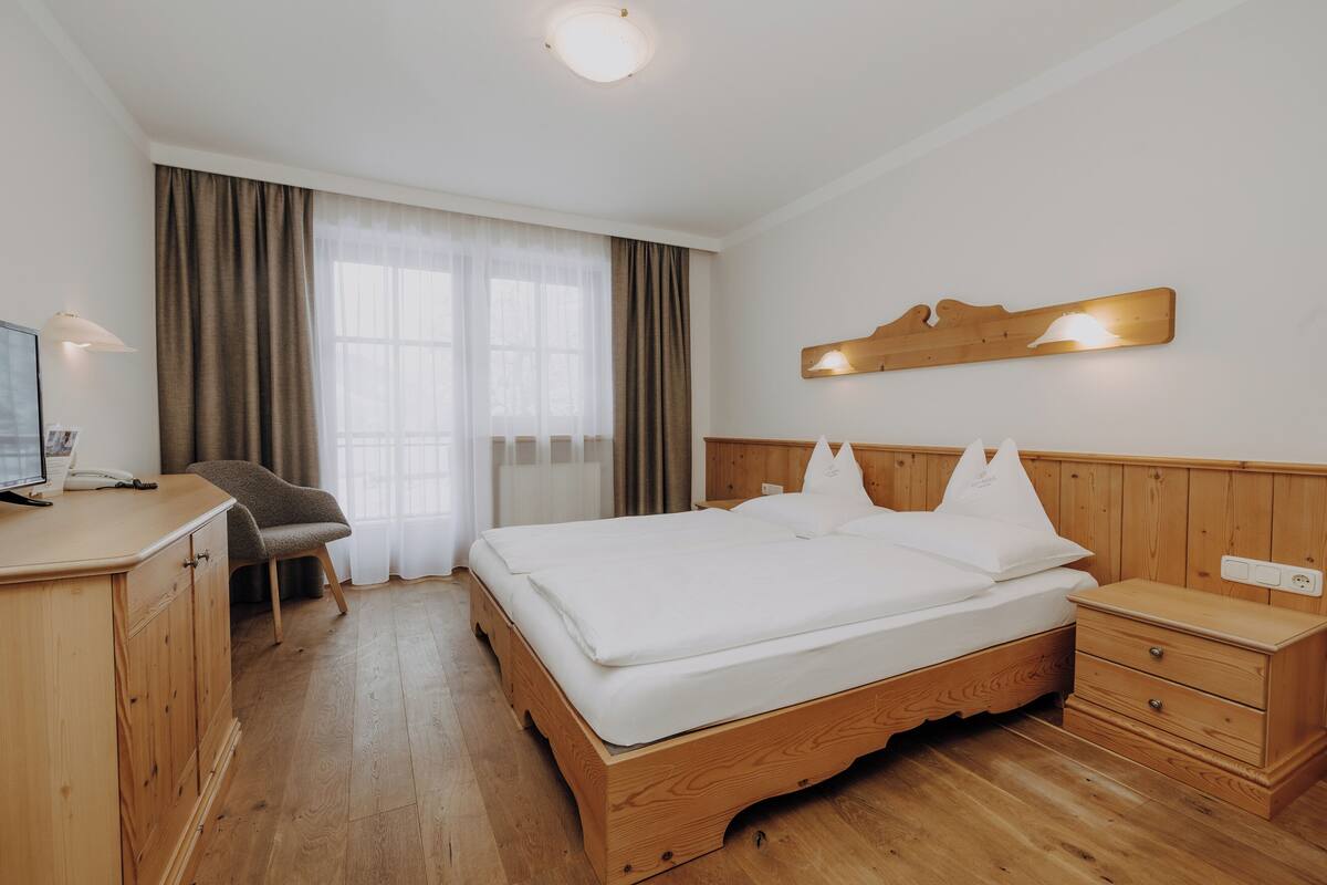 A hotel room with a wooden bed, a desk with a chair, a bedside table, and a large window with curtains.