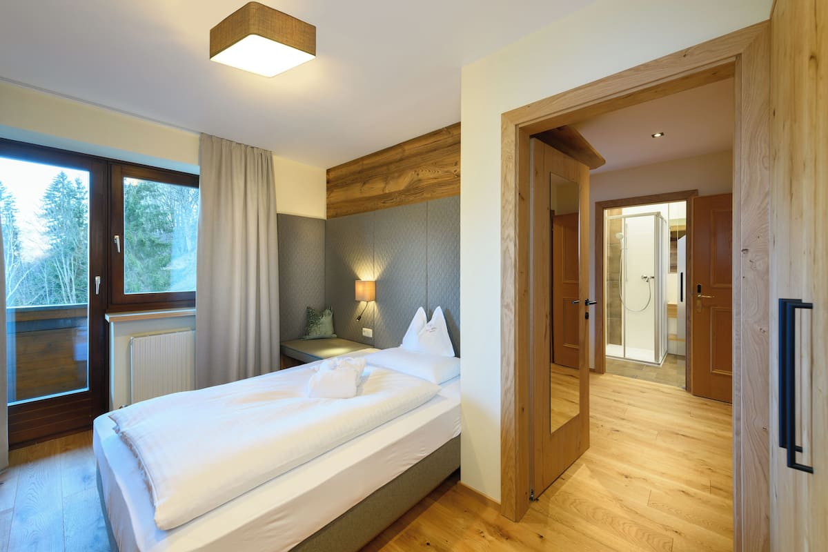 A bedroom with a bed, a window with curtains, a bedside table with a lamp, and a wooden floor.