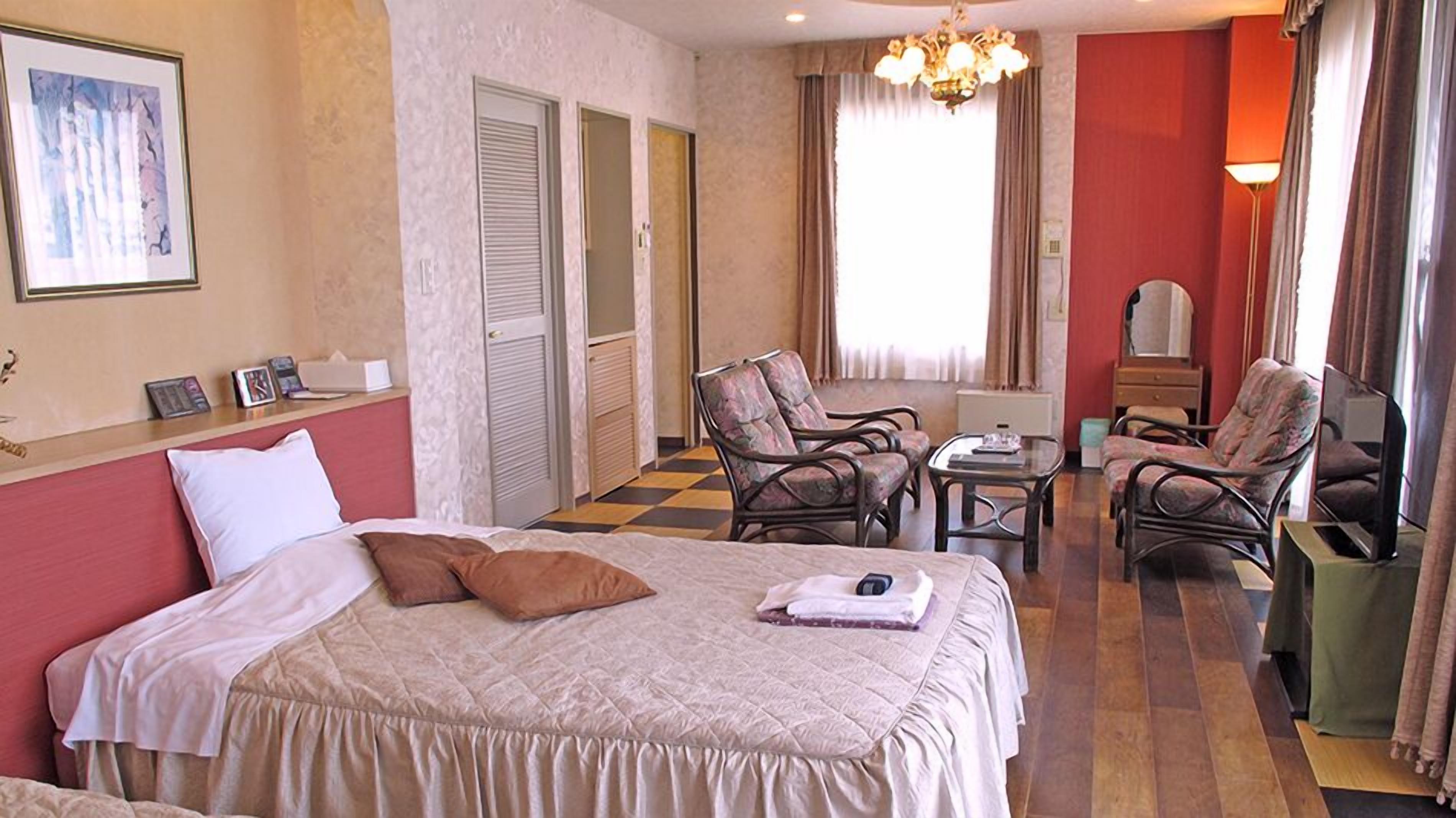 Quarto de hotel com cama, duas cadeiras, uma mesa pequena, escrivaninha e televisão.
