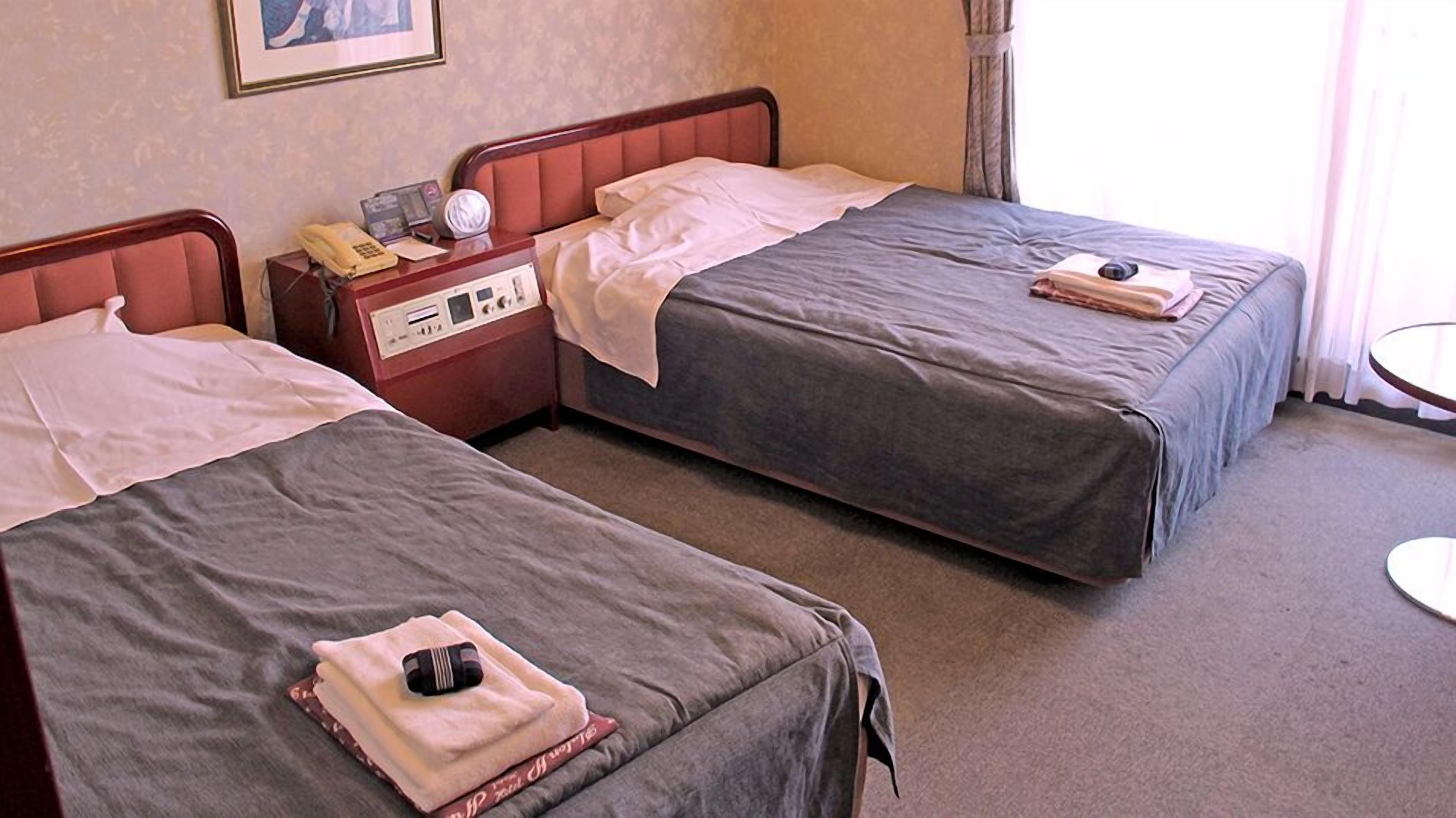 Duas camas de solteiro com lençóis cinzentos, uma mesinha de cabeceira com telefone e rádio, e uma janela com cortinas.