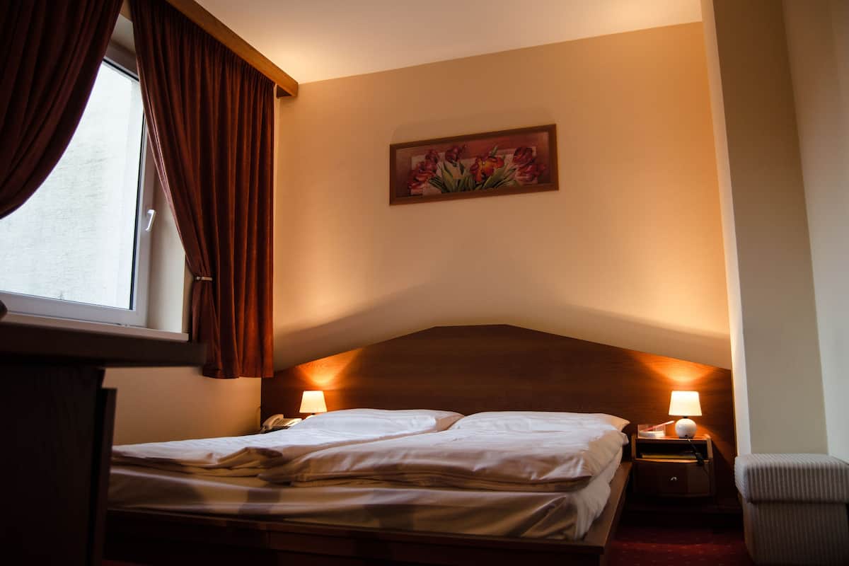 Quarto de hotel com uma cama, mesa de cabeceira com abajur, quadro na parede e janela com cortinas.