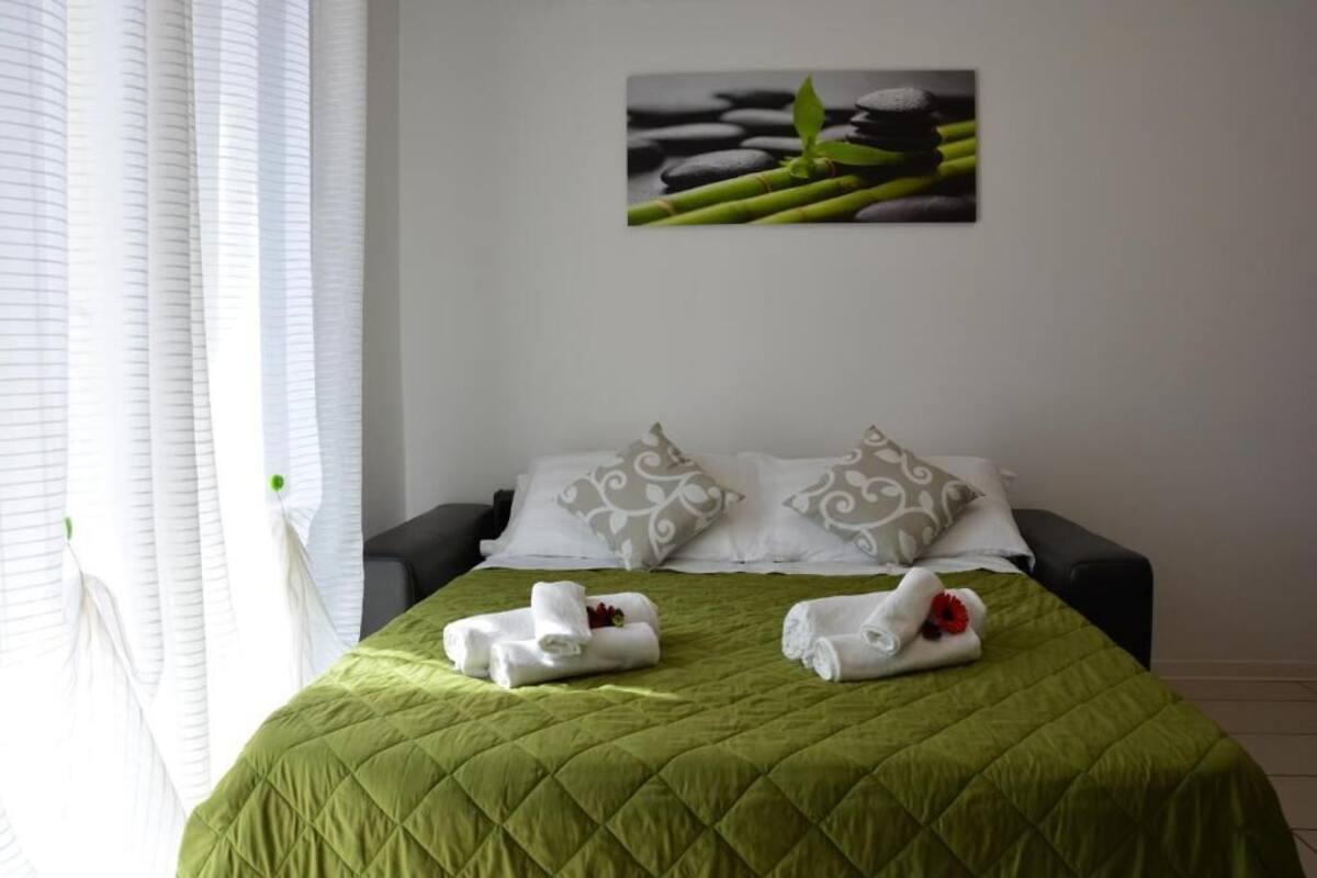 Een opgemaakt bed met een groene quilt, twee kussens met een wit en grijs patroon, en twee handdoeken met een rode bloem.