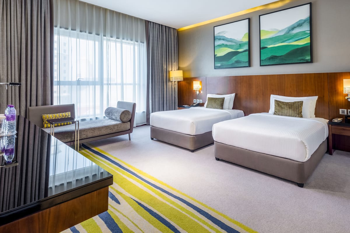 Habitación de hotel con dos camas, un escritorio, una silla y un ventanal con cortinas.