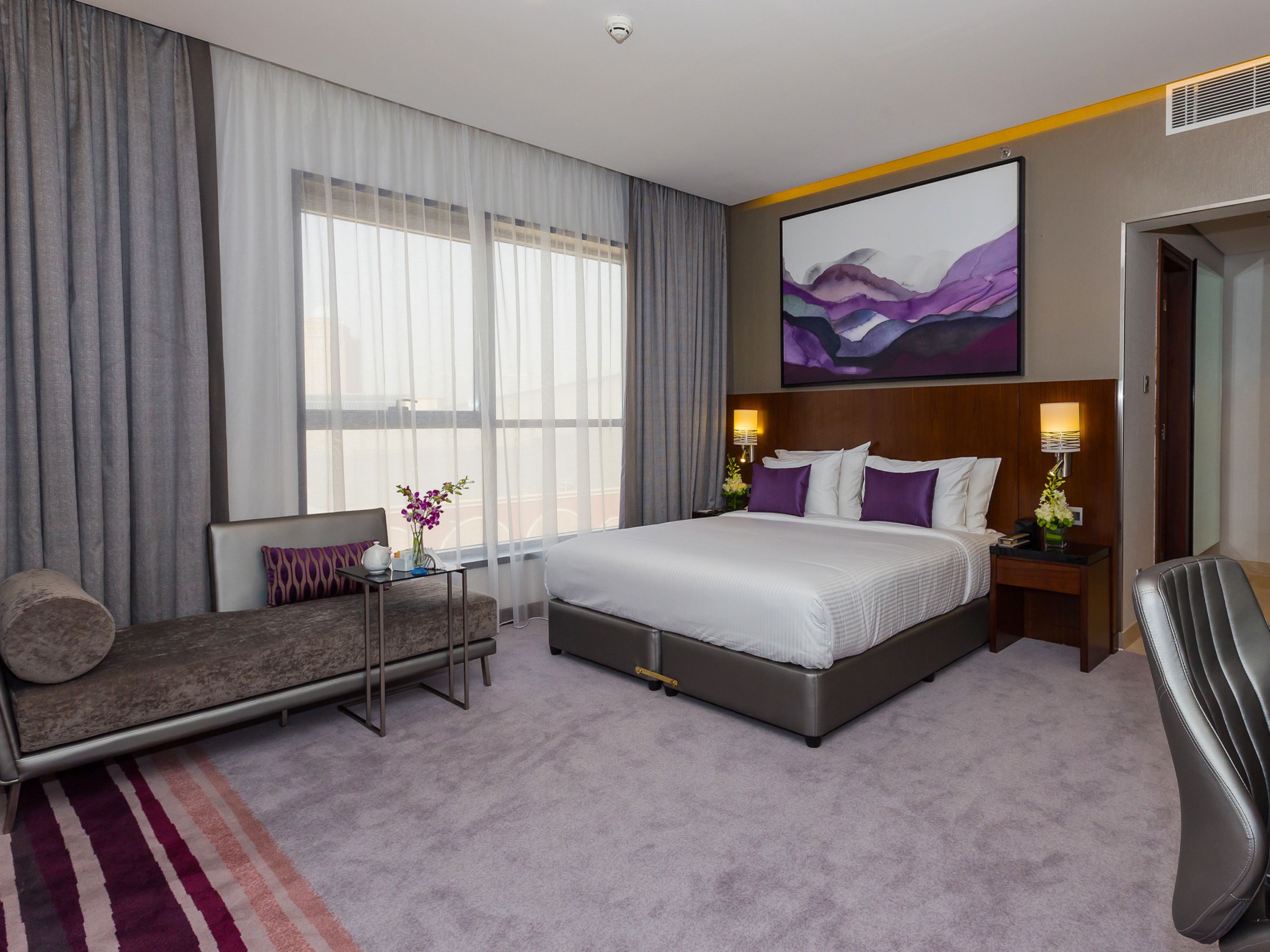 A hotel room with a large bed, a sofa, a desk, and a chair. There is a window with curtains, a painting on the wall, and a lamp on the desk.