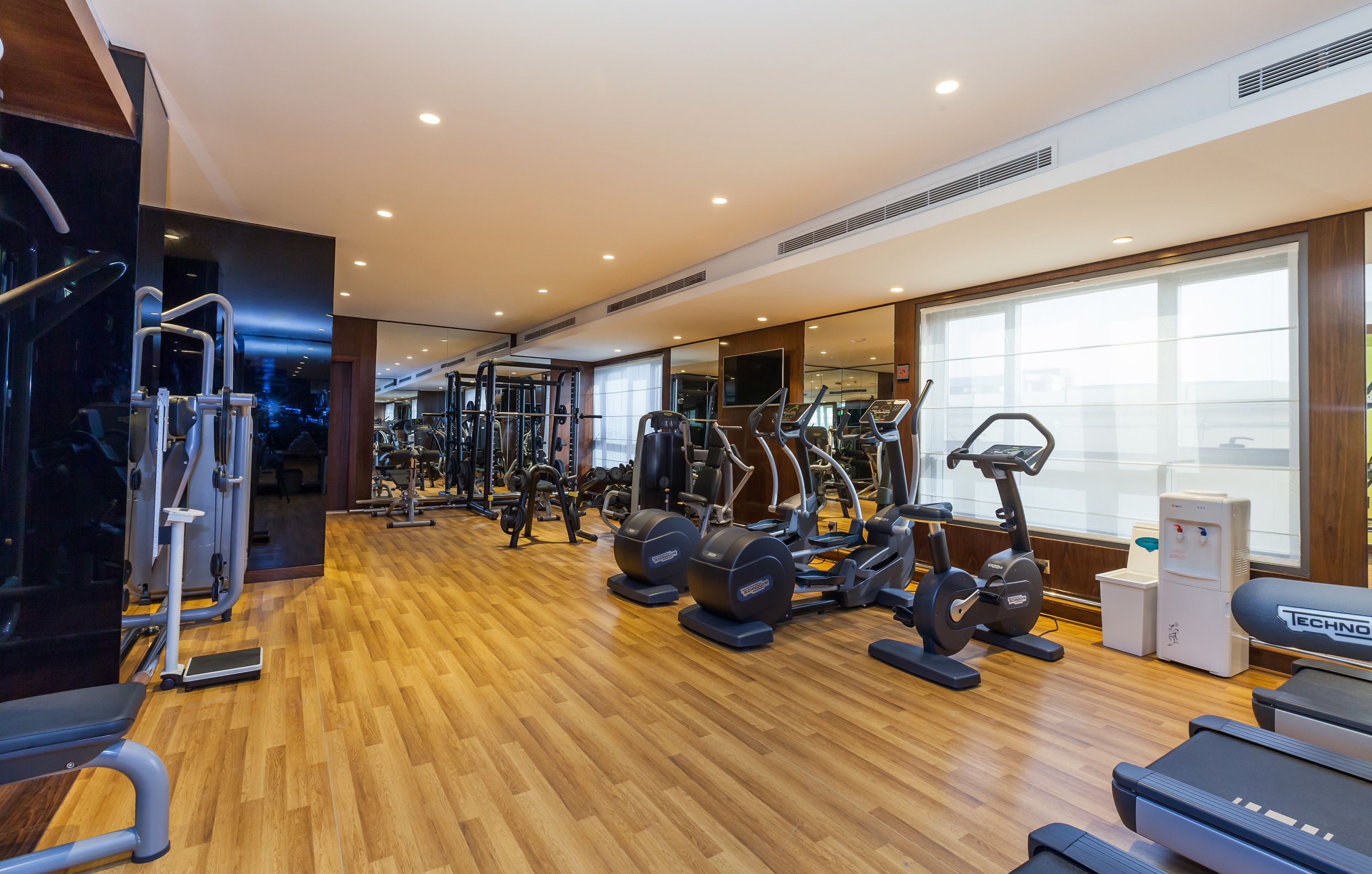 A modern gym with various exercise equipment, large windows, and a wooden floor.
