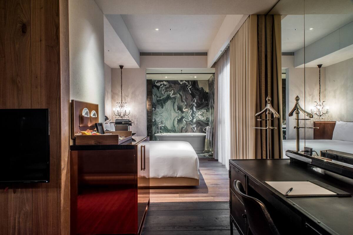 A modern hotel room with a wooden desk, a bed with a white duvet, a large mirror, and a marble wall.