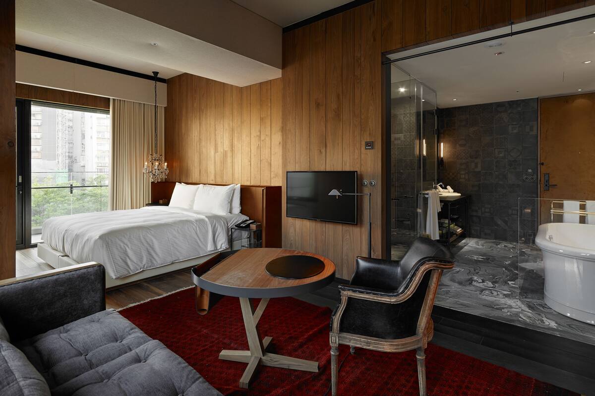 A modern hotel room with a large bed, a TV, a seating area with a table and chairs, a shower area, and a view of the city.