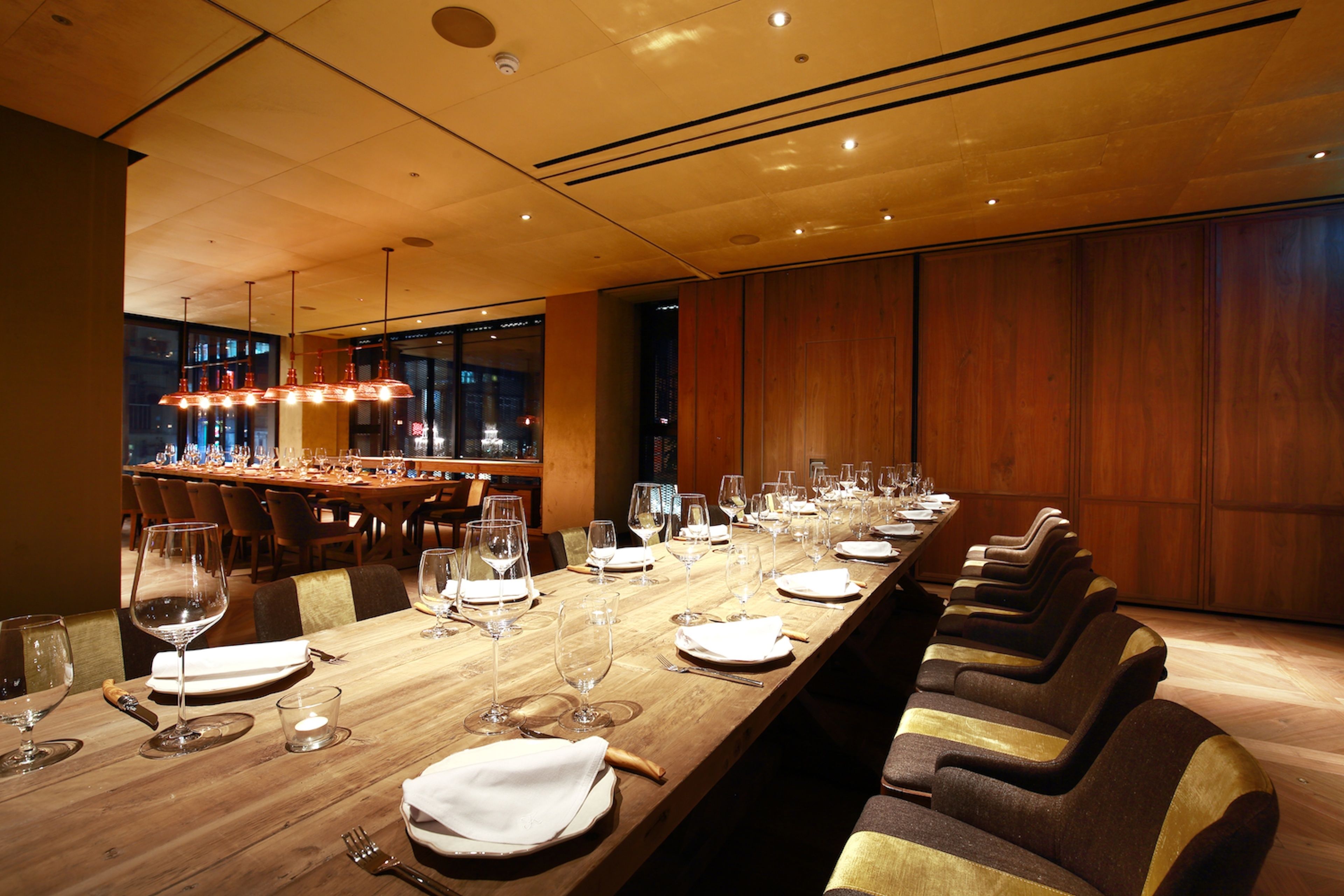 A long dining table with multiple chairs, glasses, and cutlery, set in a modern interior with wooden paneling and pendant lighting.