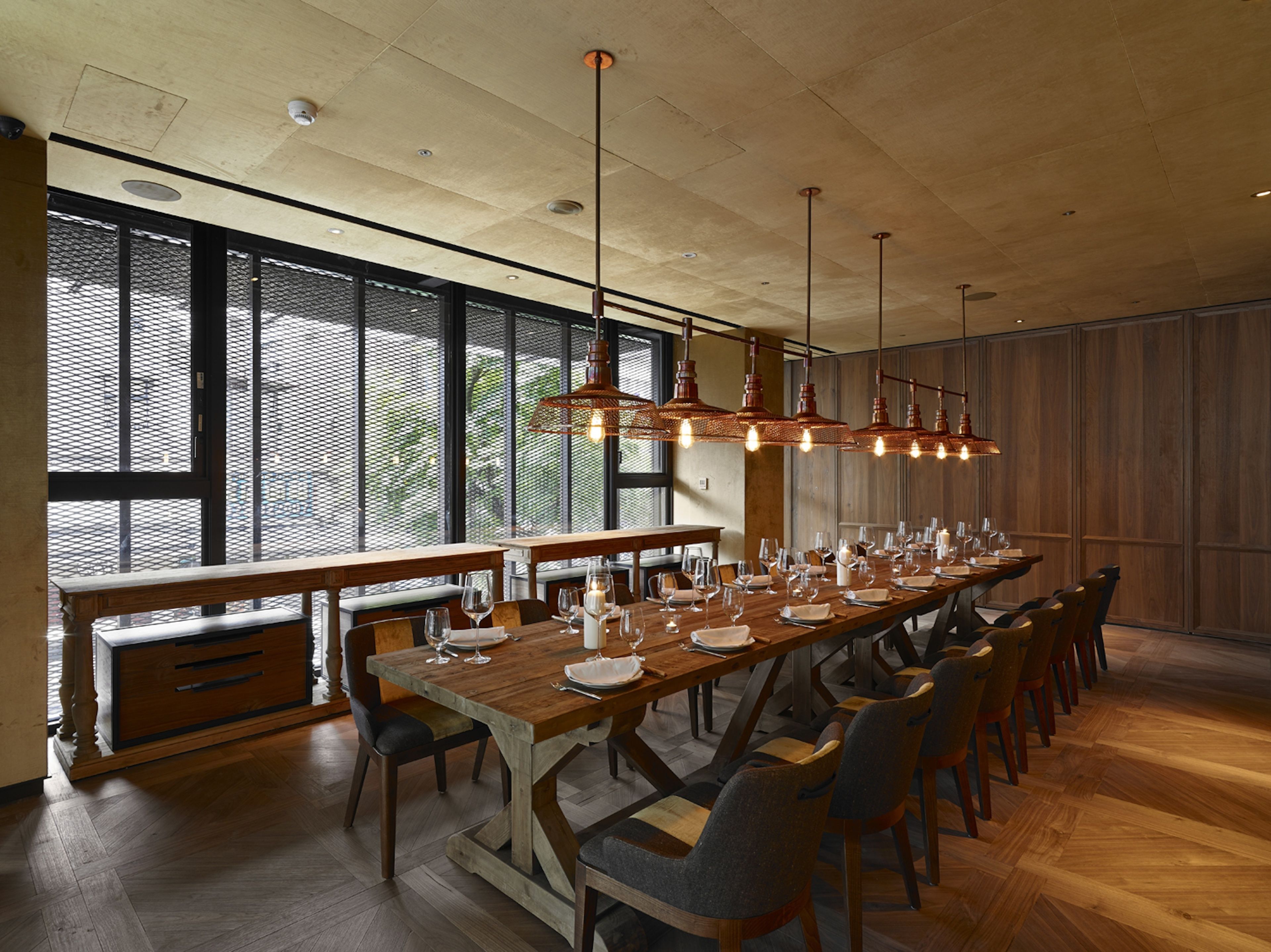 A long dining table set with chairs, wooden benches, and hanging pendant lights in a spacious room with large windows.
