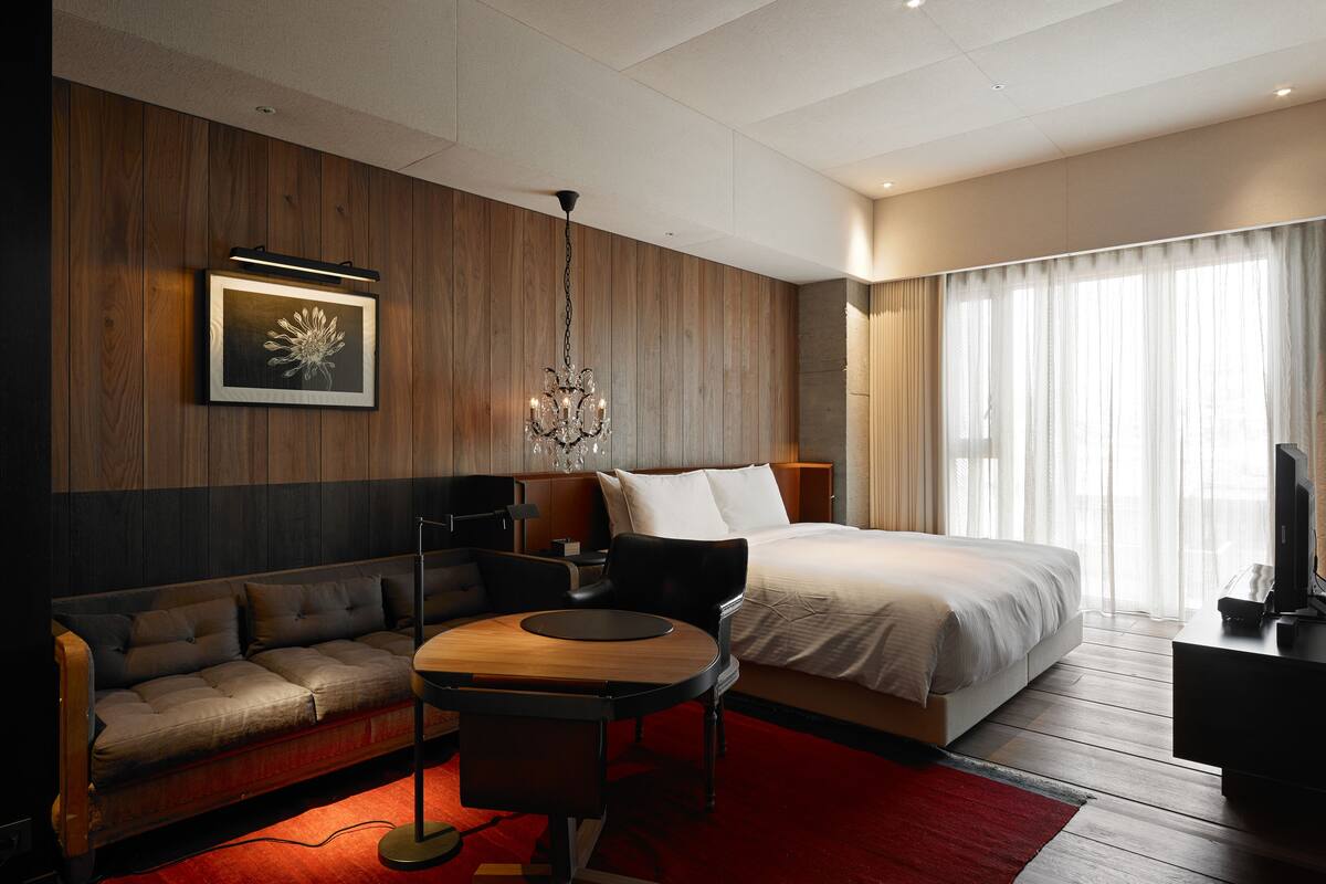 A modern hotel room with a wooden accent wall, a large bed, a sofa, a desk, and a chandelier.