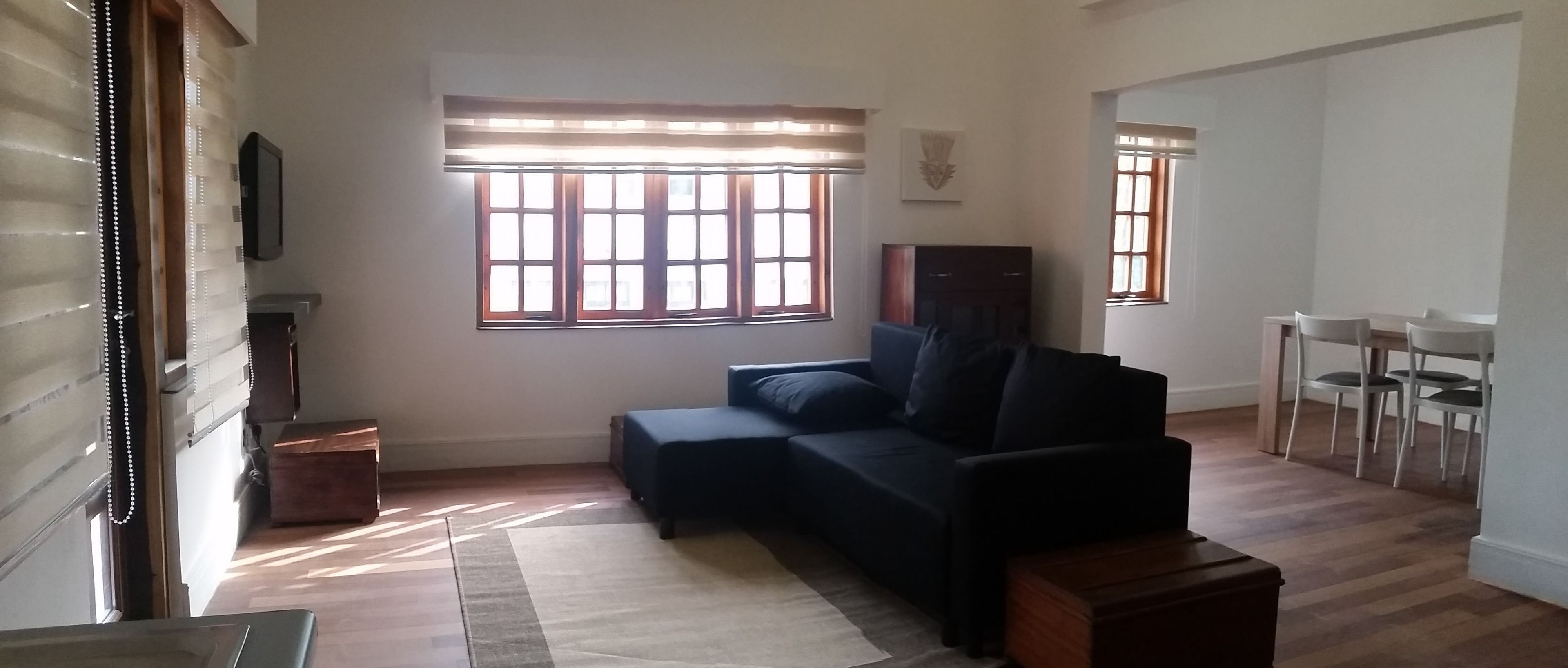 A living room with a dark-colored sofa, a wooden cabinet, and a dining area with white chairs.