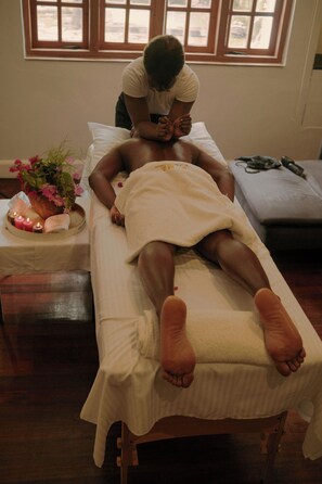 A person lying on a bed in a room with a window, a small table with candles, and a plant.
