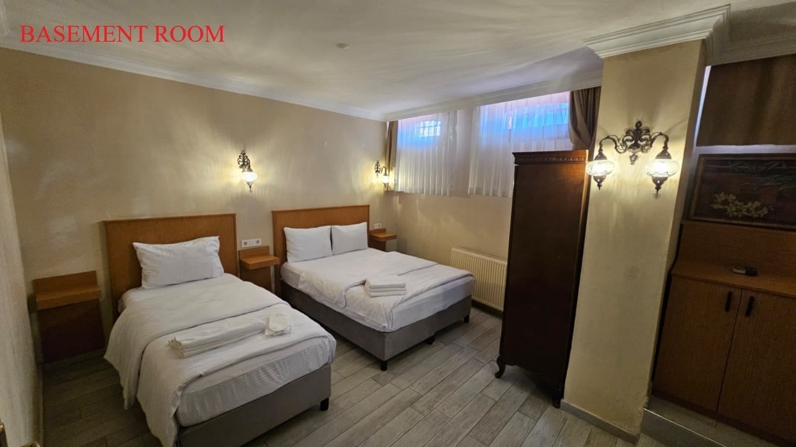 A basement room with two beds, wooden headboards, bedside tables, wall sconces, a radiator, and a wardrobe.