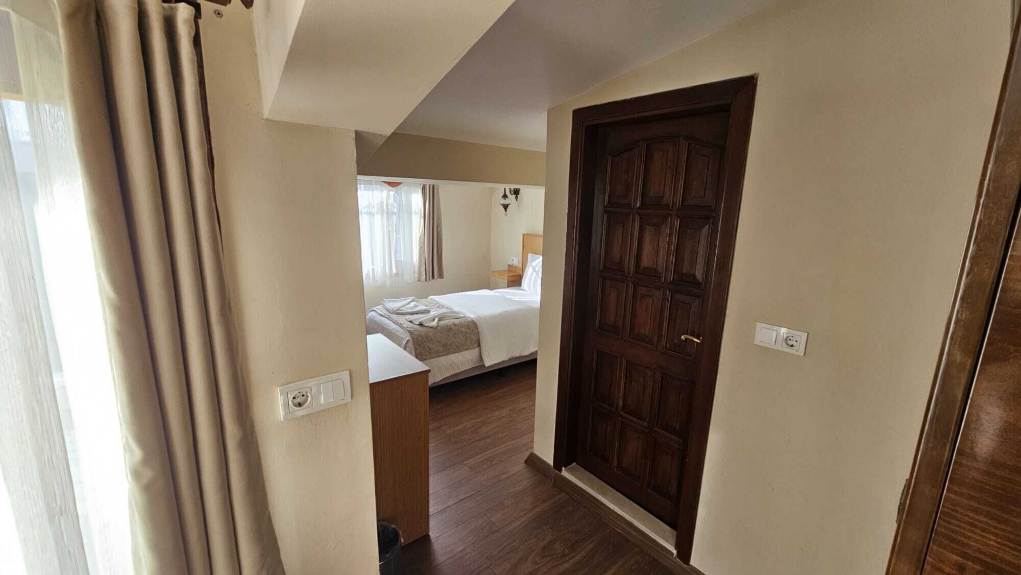 A hotel room with a large wooden door, a bed, a bedside table, and a window with curtains.