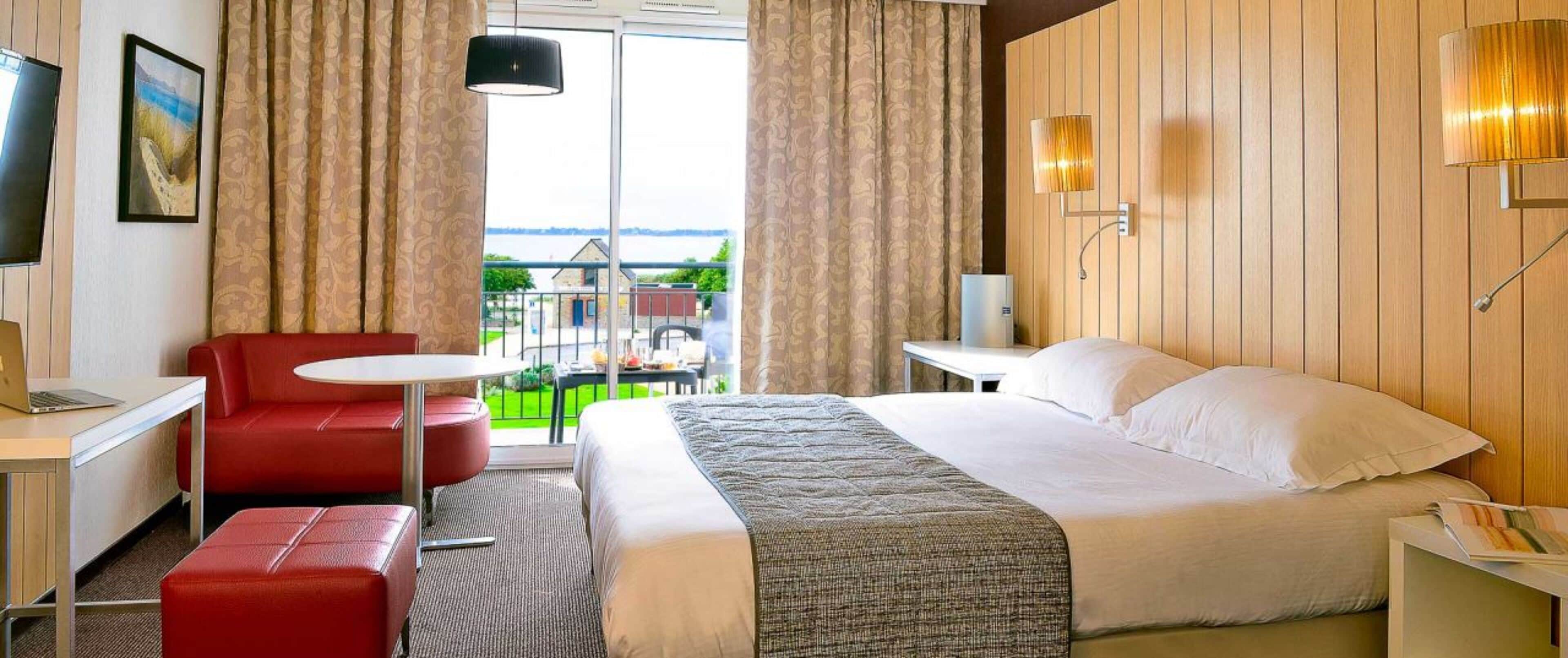 A hotel room with a bed, a desk with a laptop, a red chair, and a small table.