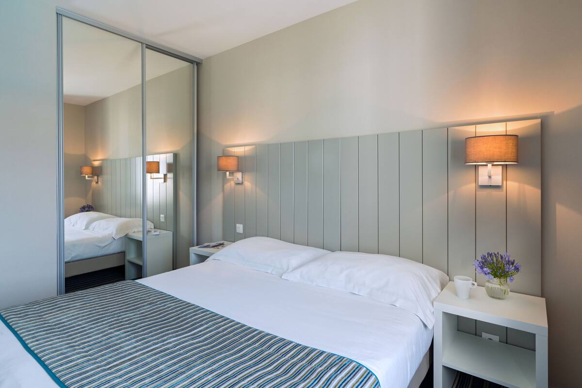 A hotel room with a large bed, a bedside table, a nightstand, a mirror, and a wall-mounted lamp.