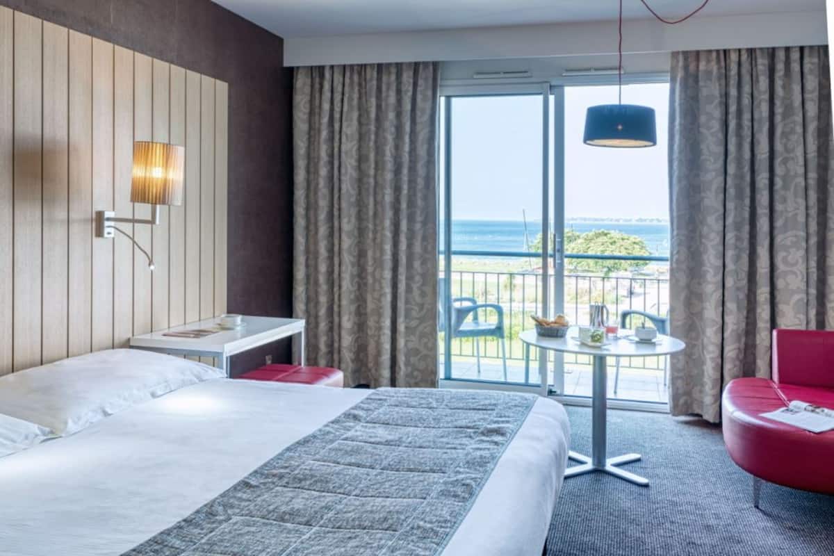A hotel room with a large bed, a small table, a red chair, and a balcony with a view of the sea.