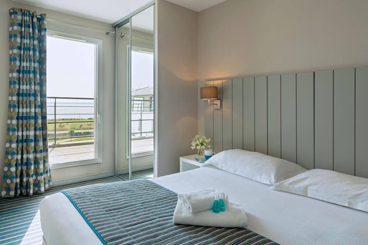 Habitación de hotel con una cama grande, una mesita de noche, una lámpara y vistas al exterior.