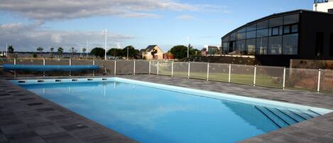 A rectangular swimming pool with a tiled deck, surrounded by a fence and a building with large windows.